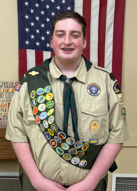 Patrick Joseph Corr III in his Scout uniform with merit-badge sash, standing in front of an American flag